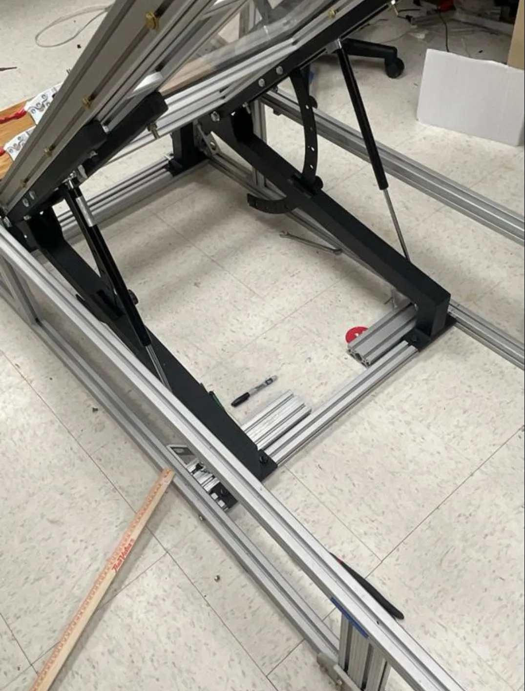 Underside of the walkway frame: black gym-bench lift mechanism, gas struts, and angle arc on a tile floor.