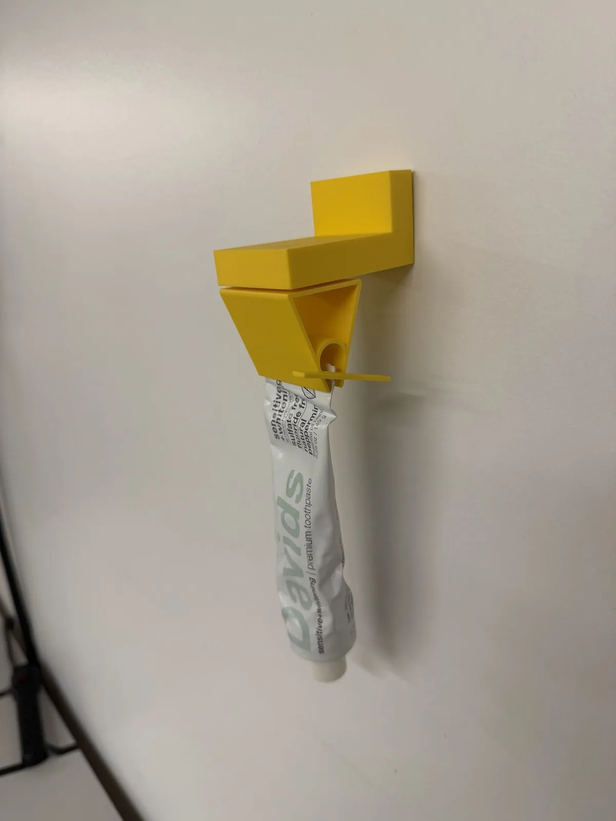Yellow 3D-printed wall dispenser on a white wall squeezing a Davids toothpaste tube through its slotted turn key.