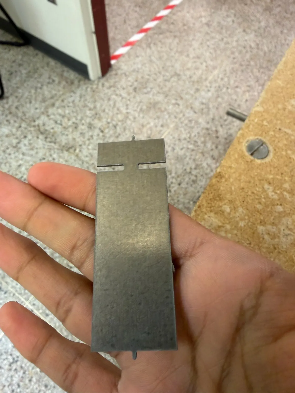 Hand holding a flat waterjet-cut steel key blank with a T-slot and small tabs, in a machine shop.